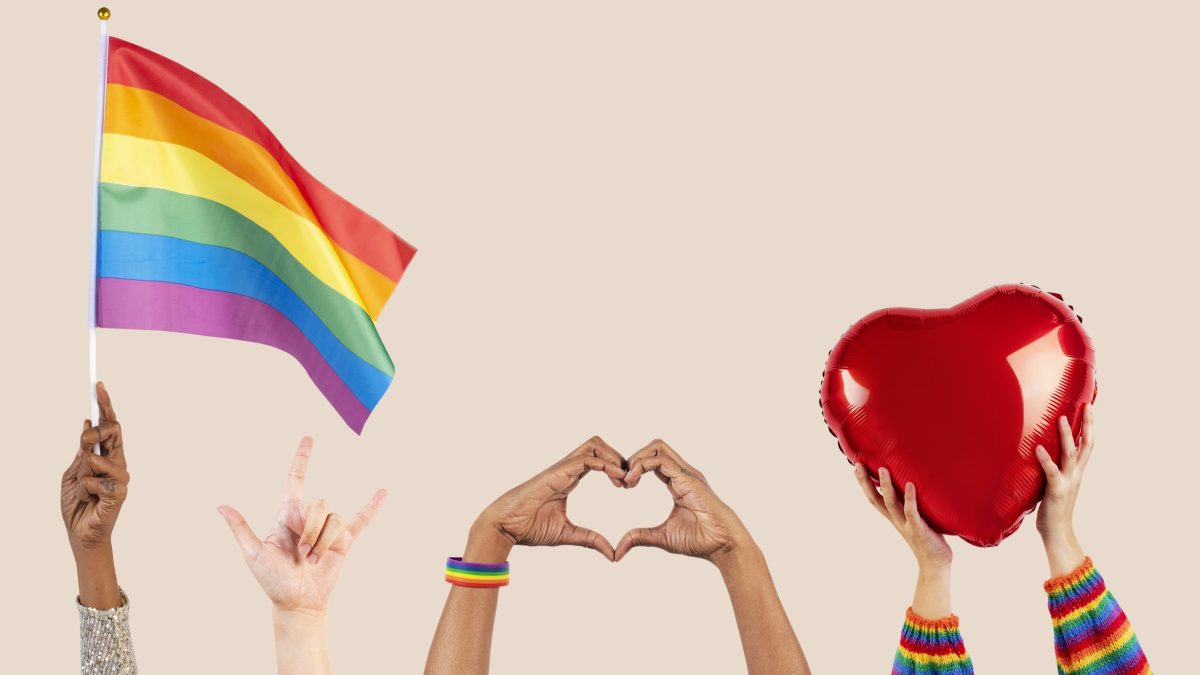 An image captures three people: one hoisting a pride flag, a second gesturing a love sign with both hands, and the third holding a red heart sign. PHOTO/Pexels