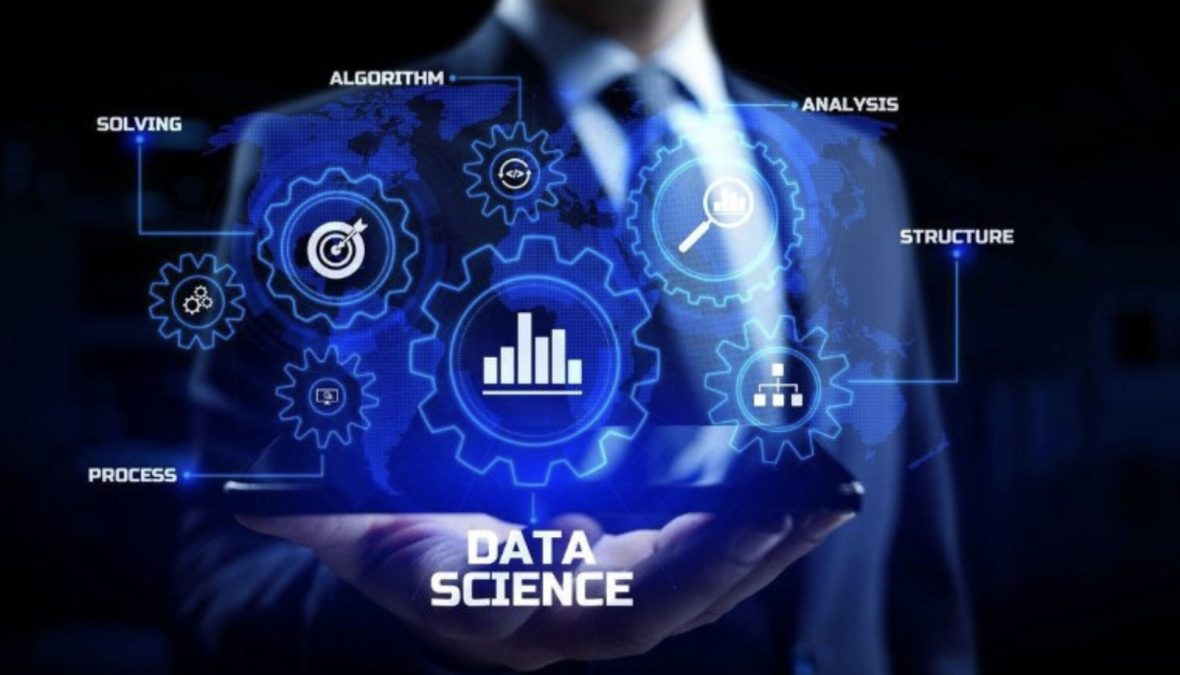 A person in a dark business suit holds a glowing blue digital interface showing interconnected gears and icons labelled “DATA SCIENCE”, with keywords such as “Algorithm”, “Analysis”, “Structure”, “Process” and “Solving” floating around the graphic. PHOTO/Pexels