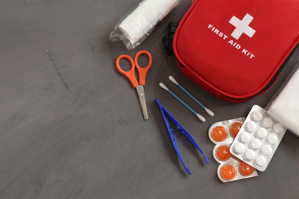 A red first aid kit placed on a grey surface alongside scissors, tweezers, cotton swabs, bandages, and blister-packed tablets. PHOTO/Pexels