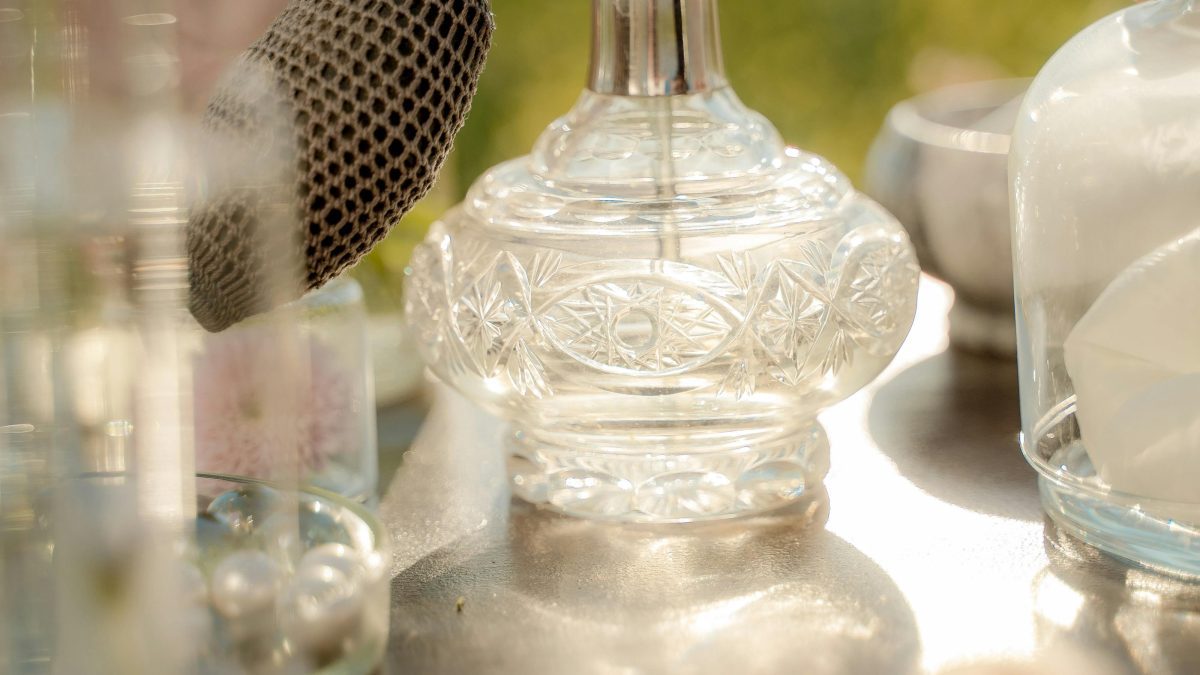 A close-up of a vintage glass perfume atomiser on a table, with soft sunlight and blurred outdoor greenery in the background. PHOTO/Pexels