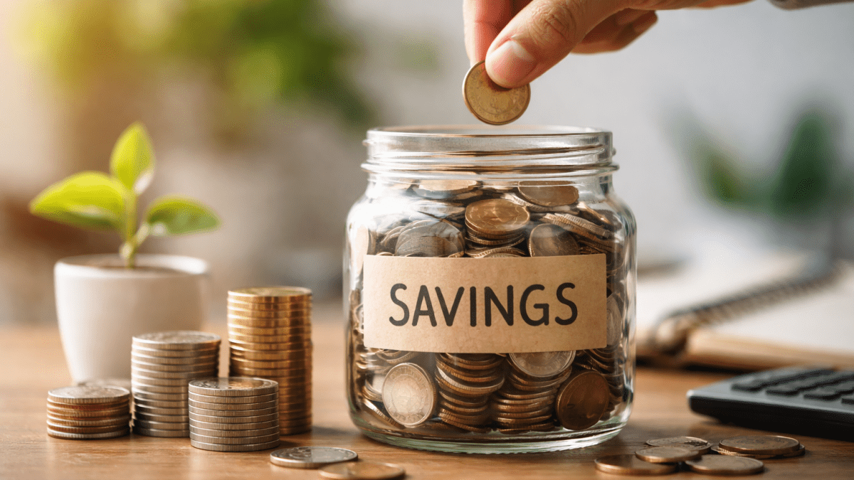 A hand drops a coin into a glass jar labeled savings, with stacks of coins placed beside it on a wooden table. PHOTO/ChatGPT/David Nthua