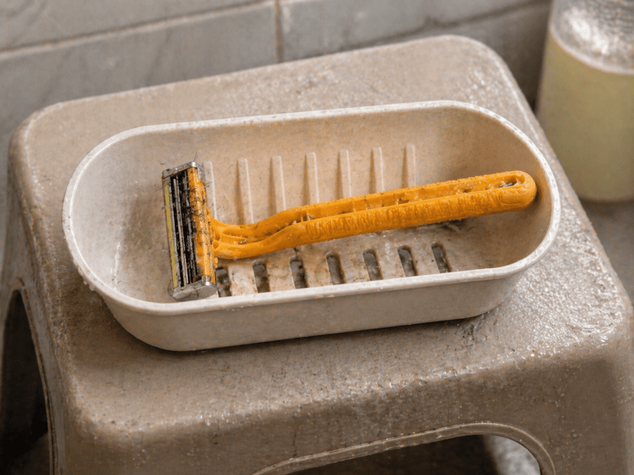 Disposable razor with hair trapped in the blades resting inside a plastic soap holder on a bathroom stool. PHOTO/Photo generated by AI
