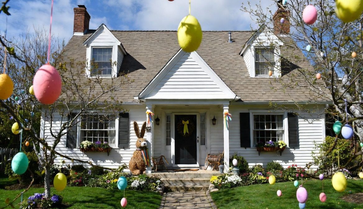 A vibrant and festive Easter landscape photo featuring a charming two-story white colonial-style house with a gray shingled roof and brick chimneys. PHOTO/Photo generated by AI