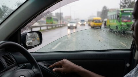 A driver's frustrated hand presses an unresponsive electric window switch during heavy rain in traffic.