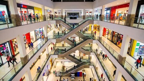 An elevated view shows the intricate, multi-level design of a major shopping mall atrium, packed with retail paths and staggered escalators under artificial light.