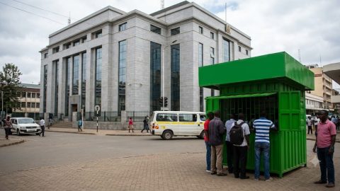 A busy mobile money kiosk operating in the shadow of a large, formal bank building.