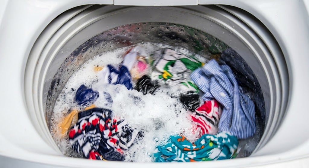 Clothes churn vigorously in the soapy water of an active top-loading washing machine during a wash cycle.