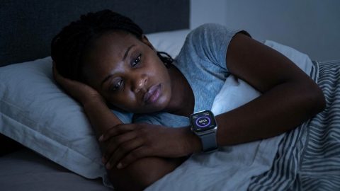 A woman in her bedroom looks anxiously at her glowing smartwatch.