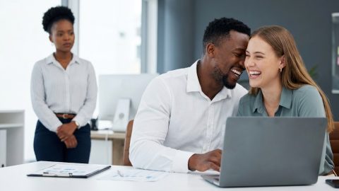 An apprehensive woman watches from a distance as her partner shares a close, laughing moment over a laptop with a female colleague in an office.